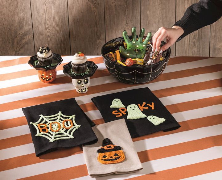 Halloween-themed decorative items including a hand-shaped basket, skull-shaped stands with cupcakes, and ghostly napkins on a striped tablecloth.