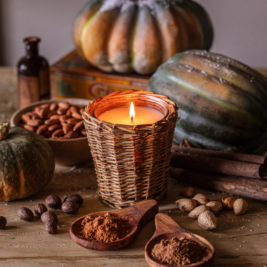 Candle in a woven holder surrounded by pumpkins, nuts, and spices on a wooden surface.