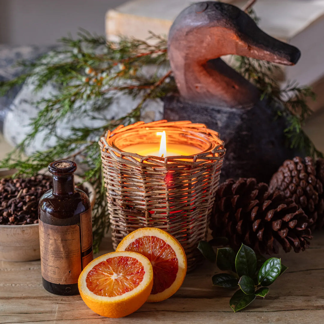Candle in a woven holder with oranges, pinecones, and a bottle on a wooden surface.